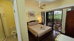 A bedroom featuring a bed with a floral arrangement, a nightstand with a lamp, and a sliding door leading to a balcony with outdoor furniture, next to a bathroom.