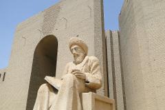 A statue of a man with a beard and turban, seated and reading a book, stands in front of a large beige brick structure with an archway, under a clear blue sky.