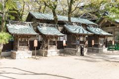 Four small wooden shrines with green tiled roofs stand in a row on a sandy ground with a building and trees in the background.