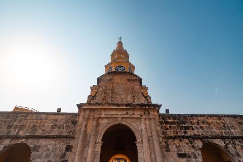 Paseo por Cartagena A low-angle shot of the Clock Tower in Cartagena, Colombia, featuring a yellow clock tower with a cross atop a weathered stone archway against a clear blue sky.