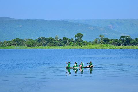 Four children in green clothing paddle a narrow boat across a calm blue lake against a backdrop of lush green trees and distant hills under a clear blue sky.