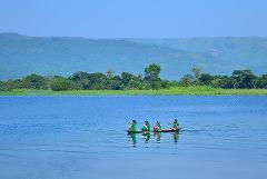 Four children in green clothing paddle a narrow boat across a calm blue lake against a backdrop of lush green trees and distant hills under a clear blue sky.