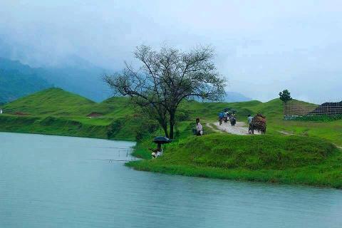 A serene landscape with rolling green hills, a lake, and people engaging in daily life, including fishing and transportation on a cloudy day.