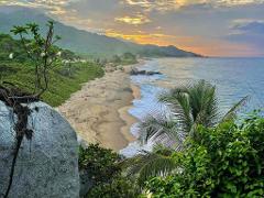 Tayrona National Park Private Luxury Tour (3 Days / 2 Nights) A scenic view of Tayrona National Park in Colombia showcases a lush green coastline meeting the ocean at sunset, with mountains in the distance and a large rock formation in the foreground.