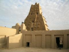 A tall mud-brick building with wooden sticks protruding from its surface rises behind a low wall with a doorway through which a woman can be seen.