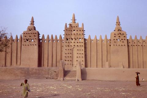 A mud-brick mosque, featuring distinctive spires and wooden sticks, stands in a sandy landscape with people nearby.