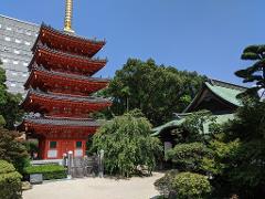 A five-story red pagoda with a golden spire stands next to a traditional Japanese building with a green roof, surrounded by lush greenery and a clear blue sky.
