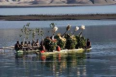 A group of people in traditional attire are on a raft sailing on water, against a backdrop of mountains.