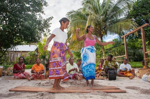 Two women dancing on wooden boards in traditional clothing in front of a group of people sitting on mats on the sand.