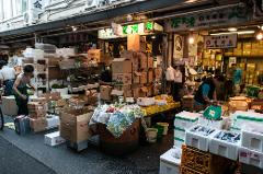 A bustling street market scene in Japan is filled with vendors selling produce and seafood from cluttered stalls overflowing with boxes and containers.
