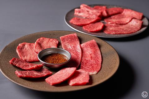 Two plates of raw, thinly sliced beef are on a gray surface, one with a small dish of sauce containing mustard seeds.