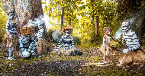 A group of people with painted faces and traditional clothing are gathered in a forest setting, some with smoke billowing around them, possibly participating in a cultural ritual.