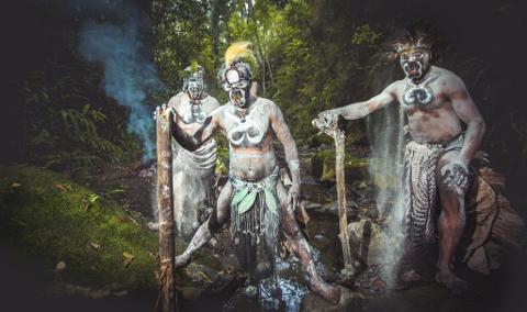 Three men in tribal attire, painted white with black facial markings, stand in a forest clearing, posing with ceremonial objects, with one man scattering powder.