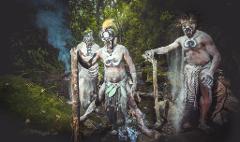 Three men in tribal attire, painted white with black facial markings, stand in a forest clearing, posing with ceremonial objects, with one man scattering powder.