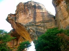 A towering rock formation with an archway is partially obscured by lush green trees.