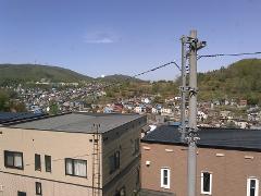 A sunlit view overlooks a hillside town with houses of various colors, nestled against green mountains under a clear blue sky.