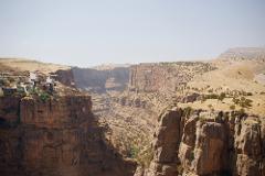 A dramatic view of a deep canyon with steep, rocky cliffs, dotted with sparse vegetation, and a small settlement perched precariously on the edge.