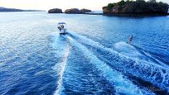 A boat tows a wakeboarder across blue water, leaving foamy trails, with rocky islands in the background.