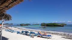 A sunny beach scene with turquoise water, rocky islands, white sand, blue lounge chairs, and a person relaxing on one of them.