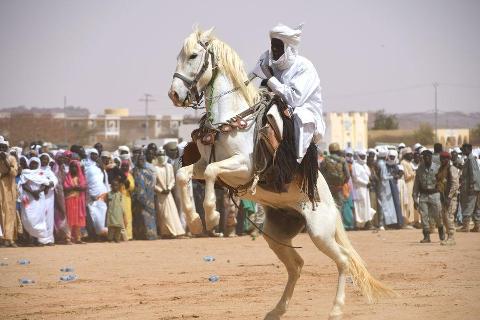 A man in white traditional clothing rides a white horse rearing up in front of a crowd of people and some military personnel on a sandy field.