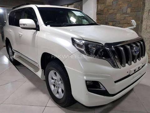 A white Toyota Land Cruiser Prado SUV is parked on a tiled surface, displaying its front fascia with a distinctive grille and headlights.