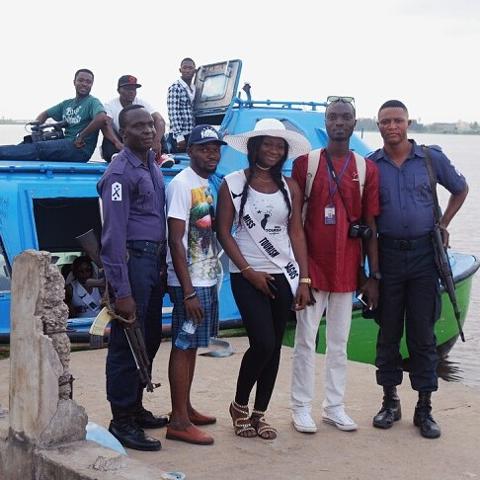 A group of people, including "Miss Tourism Lagos," pose in front of a blue boat, with two armed guards standing on either side.