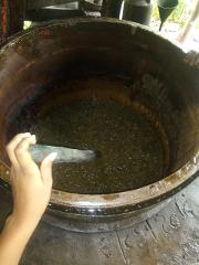 A person stirs a bubbling, dark mixture inside a large, worn-out metallic pot with a dark tool, possibly during a cooking or processing activity.