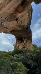 Siby Culture, History and Natural Sites Tour from Bamako A low-angle view shows a massive, weathered rock arch against a cloudy sky, with lush green trees obscuring the base.