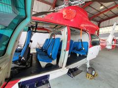 A red and white helicopter with its doors open, showing rows of blue seats inside, is parked in a hangar.