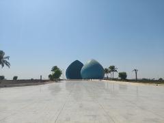 A wide view of two large, blue, droplet-shaped structures sitting on a vast, reflective paved plaza under a clear blue sky with sparse palm trees.
