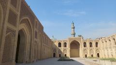 The image shows the Al-Mustansiriya Madrasah in Baghdad, Iraq, a historic building with arches, intricate brickwork, a minaret, and a central courtyard.