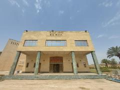 A frontal, low-angle shot of the Iraq Museum, a tan-colored building with a flat roof, large windows, and a covered entrance supported by angled columns under a blue sky.