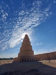 A tall, spiraling ziggurat-like structure stands prominently against a vibrant blue sky filled with scattered clouds, creating a striking architectural scene.