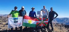A group of six people stand on a mountaintop holding flags and a sign that reads "Helgurd Mountain" with its altitude and the year 2014.