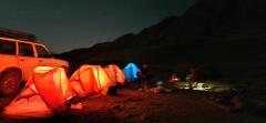 This photo shows a campsite at night with several glowing tents and a car parked nearby, set against a dark mountainous background.