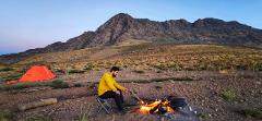 A man in a yellow jacket sits by a campfire, with an orange tent, mountains and a clear blue sky in the background.