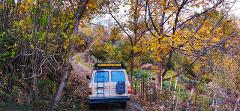 A tan-colored vehicle labeled "HALGURD MOUNTAIN GUIDE" is parked on a leaf-strewn dirt road surrounded by colorful autumn foliage in a forest.
