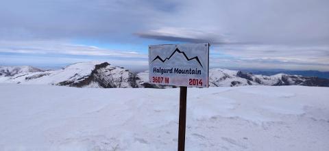 A snowy mountain landscape features a sign for Halgurd Mountain, elevation 3607m, established in 2014.