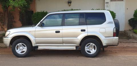 A silver Toyota Land Cruiser Prado is parked on a street next to a building with the number 318, and some greenery.