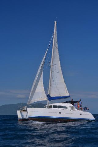 A white catamaran sails on blue water under a clear blue sky, with a few people visible on deck.