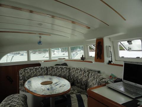 The interior of a boat's cabin features a round table surrounded by patterned seating, a laptop on a nearby surface, and windows offering a view of boats in the water.
