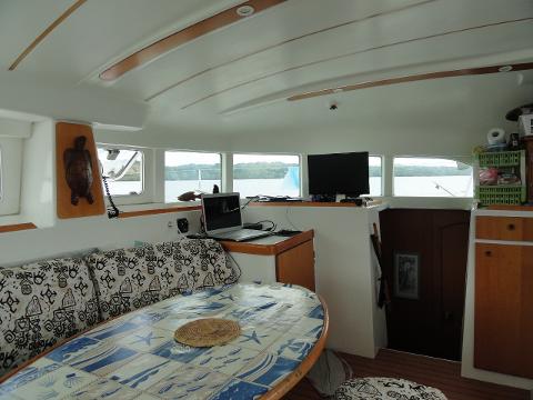 The interior of a boat features a dining area with a table, cushions, a laptop, and a TV, illuminated by natural light through the surrounding windows.
