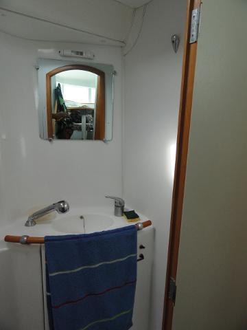 A compact boat bathroom with a white sink, faucet, and towel rack with a blue striped towel, a mirror reflecting a cabin, and a partly open wooden-framed door.