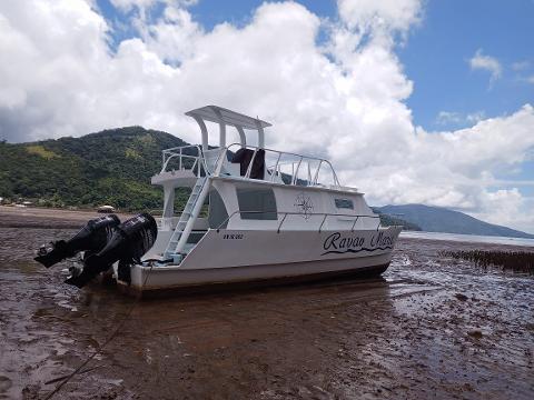 A white boat named "Ravao Maria" sits on a muddy shore under a partly cloudy sky, with green hills in the background and its two black Suzuki engines tilted upwards.