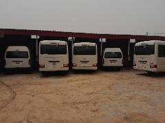Five white buses are parked under a red-roofed structure on a dirt lot, facing away from the viewer.