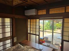 A scenic view of traditional Japanese houses with tiled roofs and lush green trees, set against a backdrop of rolling hills and a clear blue sky.