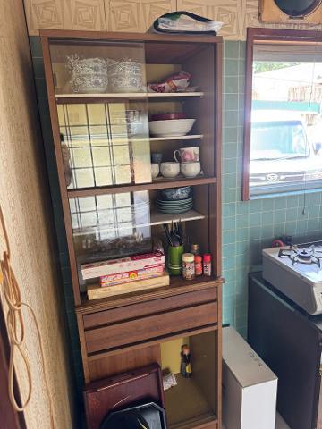 A tall wooden cabinet with glass doors displays dishes, bowls, and condiments in a kitchen setting with green tiles and a window.