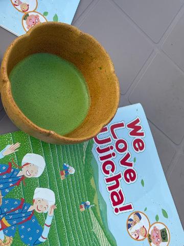 An overhead shot of a bowl of green matcha beside a "We Love Ujicha!" book on a gray surface.