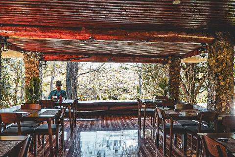 2026 - Mt. Hagen Festival Package Papua New Guinea A man sits at a table in a rustic, open-air restaurant with a wooden ceiling and stone pillars, overlooking a natural landscape.