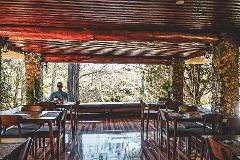 2026 - Mt. Hagen Festival Package Papua New Guinea A man sits at a table in a rustic, open-air restaurant with a wooden ceiling and stone pillars, overlooking a natural landscape.